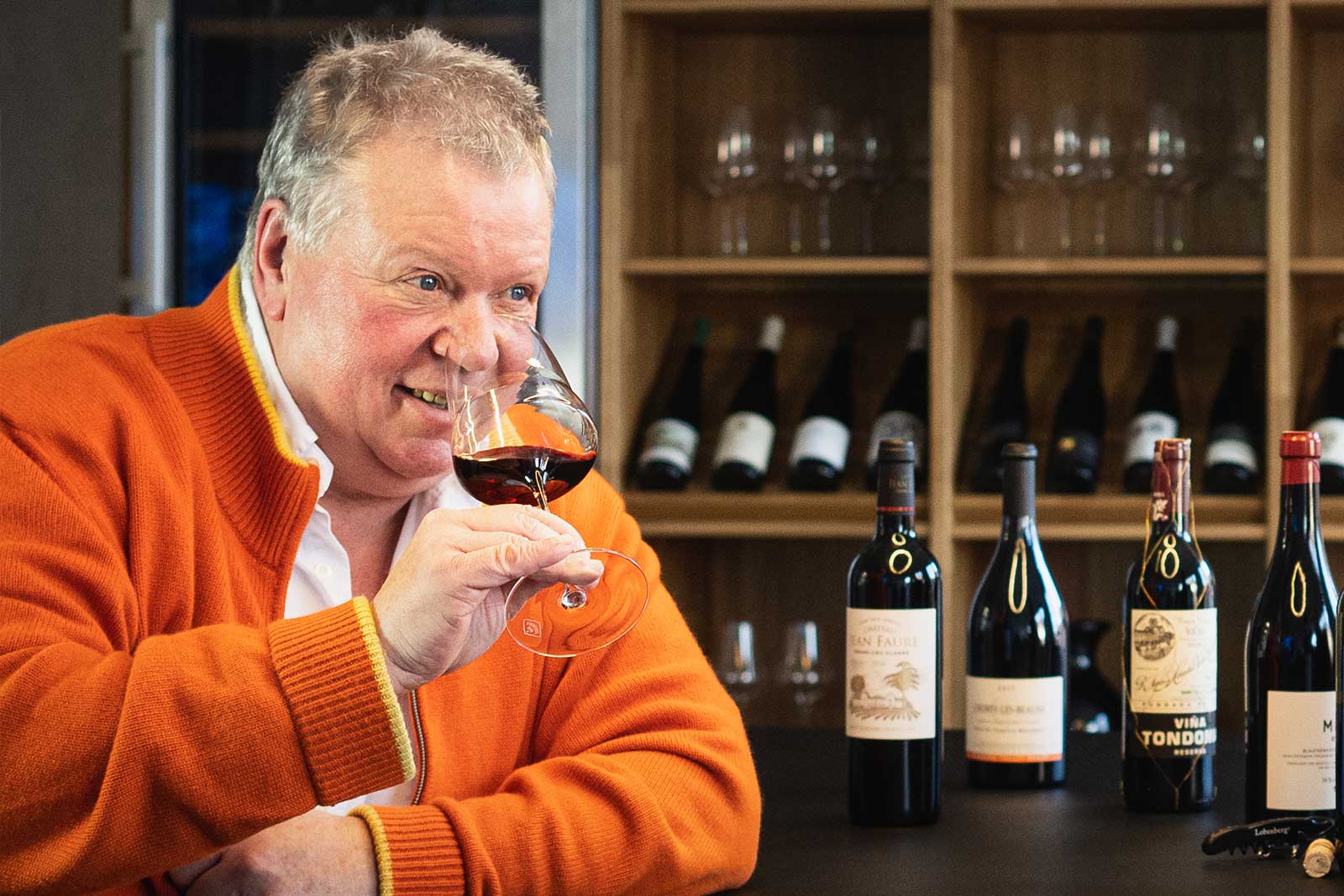 Ein älterer Mann hält ein Weinglas, das mit Rotwein gefüllt ist der Hand. Im Hintergrund stehen Weinflaschen.